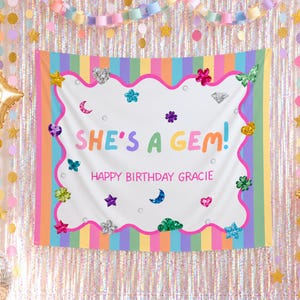 Könnte beinhalten: Ein rechteckiges Stoffbanner mit vertikalen Pastellstreifen und einer weißen Mitte mit gewelltem rosa Rand. Der Text lautet SHE'S A GEM! und HAPPY BIRTHDAY GRACIE in bunten Buchstaben, umgeben von verschiedenen Edelstein- und Sternsymbolen.