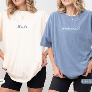 May include: Two oversized t-shirts, one ivory and one blue jean, with the words "Bride" and "Bridesmaid" embroidered on the front. The shirts have short sleeves and are paired with black shorts. The ivory shirt is on the left, and the blue jean shirt is on the right.