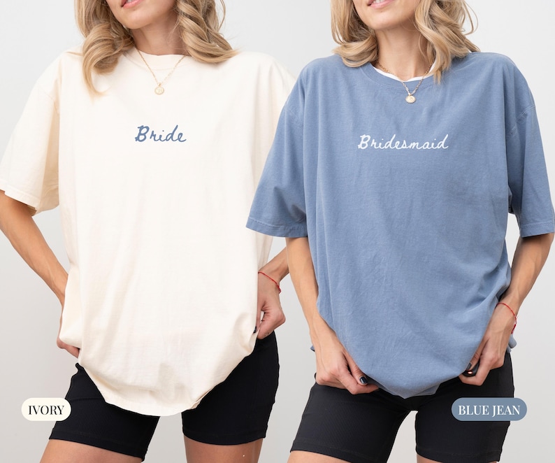 May include: Two oversized t-shirts, one ivory and one blue jean, with the words "Bride" and "Bridesmaid" embroidered on the front. The shirts have short sleeves and are paired with black shorts. The ivory shirt is on the left, and the blue jean shirt is on the right.