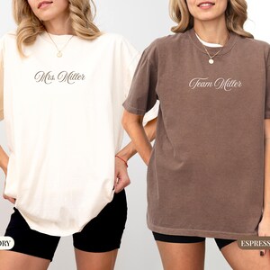 May include: Two short-sleeved t-shirts, one ivory and one espresso, with the text "Mrs. Miller" and "Team Miller" in cursive script. The shirts are worn with black shorts. The ivory shirt has a tag that says "IVORY" and the espresso shirt has a tag that says "ESPRESSO".
