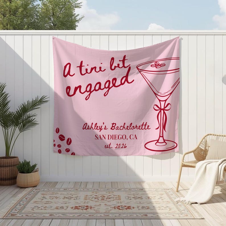 K&ouml;nnte beinhalten: Ein rosa Stoffbanner mit dem Text "a tini bit engaged" in roter Schrift. Rechts befindet sich eine rote Martini-Glas-Grafik. Darunter steht "Ashley's Bachelorette, San Diego, CA, est. 2026".
