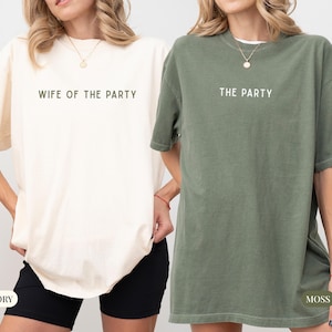 May include: Two oversized t-shirts, one ivory and one moss green, are displayed. The ivory shirt reads "WIFE OF THE PARTY" in a simple font. The moss green shirt reads "THE PARTY". Both shirts have short sleeves and a relaxed fit.