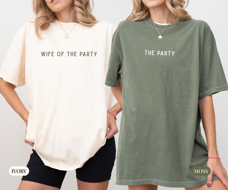 May include: Two oversized t-shirts, one ivory and one moss green, are displayed. The ivory shirt reads "WIFE OF THE PARTY" in a simple font. The moss green shirt reads "THE PARTY". Both shirts have short sleeves and a relaxed fit.