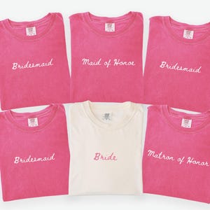 May include: A collection of pink t-shirts with white embroidered text, including "Bridesmaid", "Maid of Honor", and "Matron of Honor". A single off-white shirt is embroidered with "Bride". The shirts are likely for a wedding party.