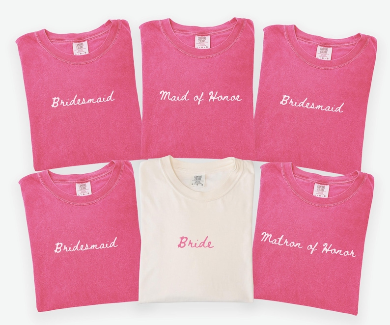 May include: A collection of pink t-shirts with white embroidered text, including "Bridesmaid", "Maid of Honor", and "Matron of Honor". A single off-white shirt is embroidered with "Bride". The shirts are likely for a wedding party.