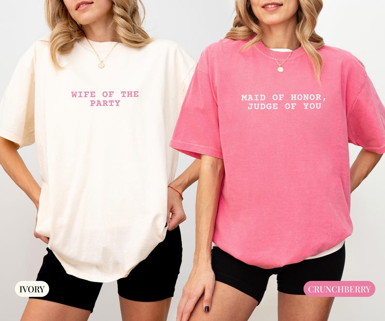 May include: Two oversized t-shirts, one ivory and one crunchberry pink, with text. The ivory shirt reads "WIFE OF THE PARTY" in pink lettering. The pink shirt says "MAID OF HONOR, JUDGE OF YOU" in white lettering. Both shirts are paired with black shorts.