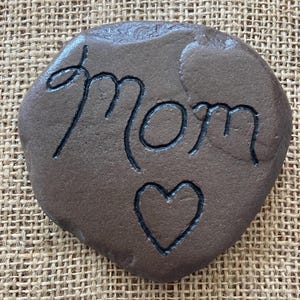 May include: A brown stone with the word "Mom" and a heart carved into it.