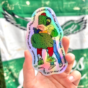 May include: A holographic sticker with a cartoon illustration of two people hugging. The sticker has the text "PHILADELPHIA NO ONE LIKES US. WE DON'T CARE" repeated around the edge.