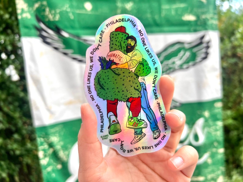 May include: A holographic sticker with a cartoon illustration of two people hugging. The sticker has the text "PHILADELPHIA NO ONE LIKES US. WE DON'T CARE" repeated around the edge.