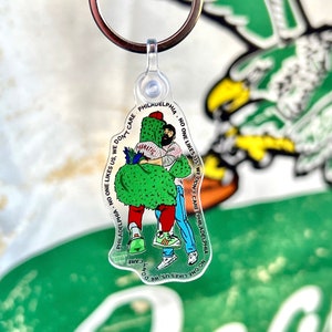May include: A clear plastic keychain with a green and red cartoon character hugging a person. The text on the keychain reads "PHILADELPHIA - NO ONE LIKES US. WE DON'T CARE. PHILADELPHIA - NO ONE LIKES US. WE DON'T CARE."