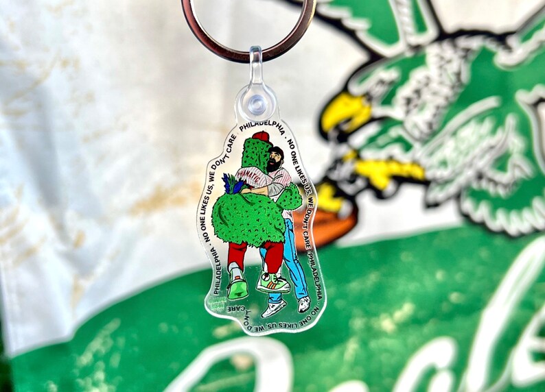May include: A clear plastic keychain with a green and red cartoon character hugging a person. The text on the keychain reads "PHILADELPHIA - NO ONE LIKES US. WE DON'T CARE. PHILADELPHIA - NO ONE LIKES US. WE DON'T CARE."