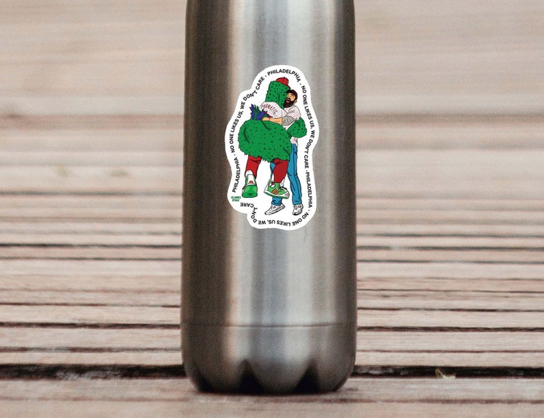 May include: A white sticker with a green, cartoon cactus hugging a person. The sticker has text that reads "NO ONE LIKES US WE DON'T CARE" PHILADELPHIA, NO ONE LIKES US WE DON'T CARE" PHILADELPHIA.