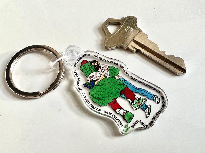 May include: A white acrylic keychain with a cartoon illustration of two people. The keychain features the text "Philadelphia - No One Likes Us, We Don't Care - Philadelphia - No One Likes Us, We Don't Care".