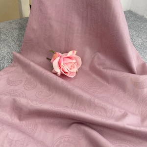 Pode incluir: Um tecido rosa claro com um padrão paisley. O tecido está drapeado sobre uma superfície e uma rosa rosa é colocada em cima do tecido.