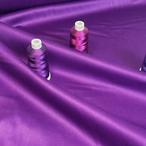 May include: A close-up shot of vibrant purple satin fabric with a subtle sheen. Several spools of purple thread are scattered across the fabric, adding texture and color contrast. The fabric is draped, creating soft folds and shadows.