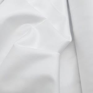 Op de afbeelding: Close-up van witte stof met zachte plooien en subtiele schaduwen. De stof lijkt glad en mogelijk satijn of zijde te zijn, met een lichte glans. De afbeelding benadrukt de textuur en de drapering van het materiaal.