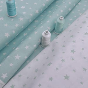 Peut inclure: Plusieurs rouleaux de tissu à motifs d'étoiles bleues et blanches sont présentés. Trois bobines de fil, deux bleues et une blanche, sont posées sur le tissu. Le tissu est probablement destiné à des projets de couture ou d'artisanat.
