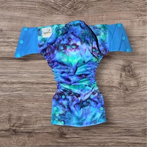 May include: A reusable cloth diaper with a vibrant tie-dye pattern in shades of blue, green, and purple. The diaper has blue wings with snap closures and a tag that reads "Squishie". The diaper is designed for babies and toddlers.