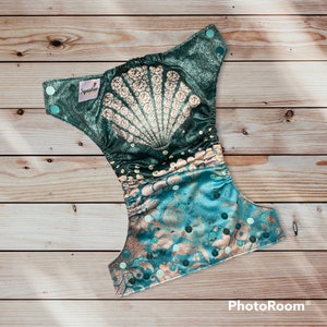 May include: A teal and green reusable cloth diaper with a large seashell design and a smaller seashell pattern on the bottom. The diaper has a snap closure and is made by the brand "Squishbee".