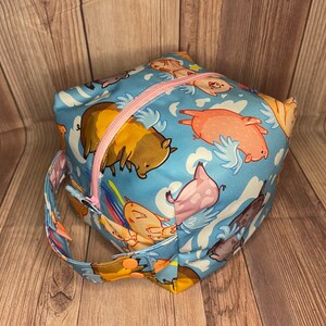 May include: A blue fabric cube-shaped bag with a pink zipper and a handle. The bag has a pattern of cartoon pigs with wings flying in the clouds.