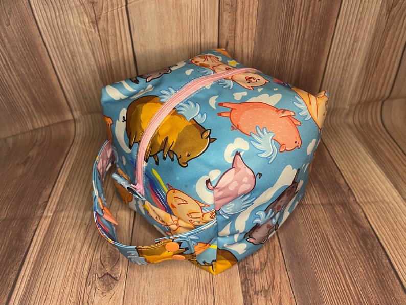 May include: A blue fabric cube-shaped bag with a pink zipper and a handle. The bag has a pattern of cartoon pigs with wings flying in the clouds.