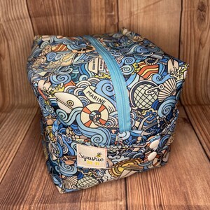 May include: A blue and yellow zippered pouch with a nautical theme. The pouch has a variety of illustrations including anchors, boats, waves, and the word "Marine". The pouch has a white label with the text "Squishie Bee-By" and a bee illustration.