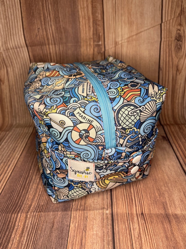 May include: A blue and yellow zippered pouch with a nautical theme. The pouch has a variety of illustrations including anchors, boats, waves, and the word "Marine". The pouch has a white label with the text "Squishie Bee-By" and a bee illustration.