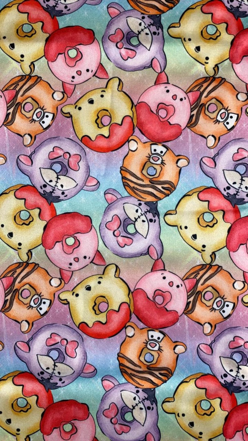 May include: A colorful fabric with a repeating pattern of cartoon donuts. Each donut has a different character face on it, including a bear, a tiger, and a pig. The donuts are all pink, yellow, or purple and have red icing.