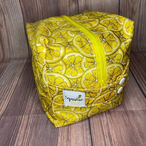May include: Yellow zippered pouch with a lemon slice pattern and a white button. The pouch has a label that says "Squishie Bee-By".