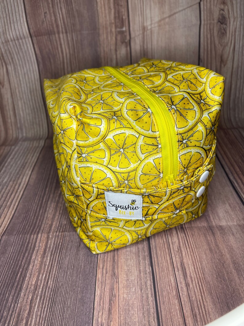 May include: Yellow zippered pouch with a lemon slice pattern and a white button. The pouch has a label that says "Squishie Bee-By".