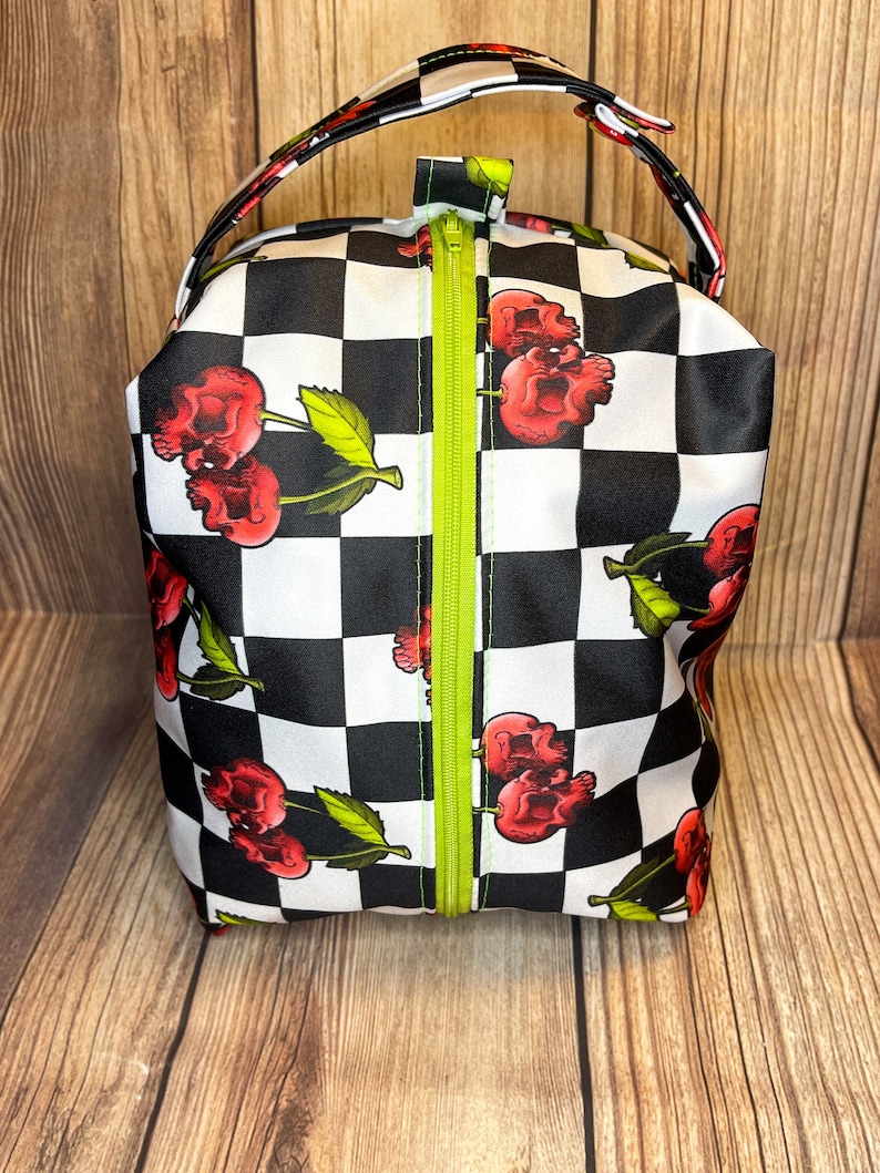 May include: A black and white checkered zippered pouch with a skull and cherry design. The pouch has a green zipper and a black handle.