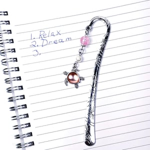 May include: A silver metal bookmark with a decorative design and a dangling charm. The charm features a turtle pendant with a pink center, a pink bead, and a pearl. The words "1. Relax 2. Dream" are written on the lined paper.