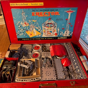 May include: Vintage Erector Set No. 10 in a red metal case. The lid features a colorful illustration of amusement park rides. Inside, various metal construction pieces, gears, and tools are neatly organized, offering a glimpse into classic engineering and building toys.