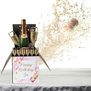 May include: A pop-up card with a black box and a champagne bottle with glasses inside. The card has a pink and gold watercolour background with balloons and the text "Happy Birthday Jo".