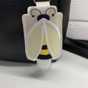 May include: A white hand sanitiser holder with a bee design. The bee is embroidered with black, yellow and white thread. The holder is attached to a silver keyring.