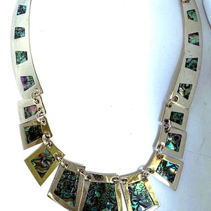 May include: A gold-toned metal necklace featuring a series of connected, trapezoidal links. Each link is inlaid with iridescent abalone shell segments displaying vibrant green, blue, and purple hues. The necklace has a curved, collar-like shape.