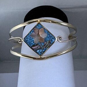 May include: A gold-toned cuff bracelet featuring a central diamond-shaped inlay. The inlay displays a light pink mother-of-pearl flower set against a background of crushed blue turquoise-colored stone, with a twisted metal wire detail on the sides.