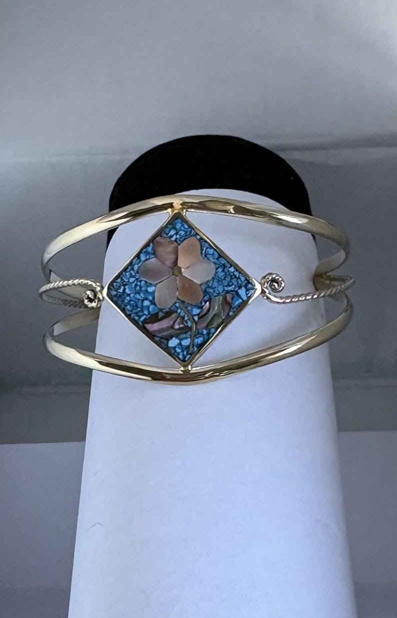 May include: A gold-toned cuff bracelet featuring a central diamond-shaped inlay. The inlay displays a light pink mother-of-pearl flower set against a background of crushed blue turquoise-colored stone, with a twisted metal wire detail on the sides.