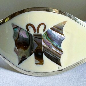 May include: A wide cuff bracelet featuring a central butterfly inlay made of iridescent abalone shell set against a smooth cream-colored background. The bracelet is framed by a polished silver-toned metal border and includes additional abalone accents.