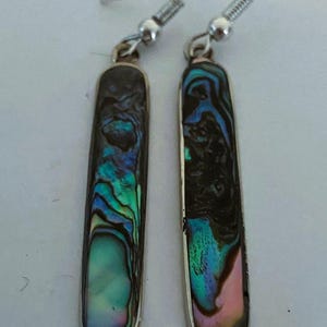 May include: A pair of rectangular dangle earrings with iridescent abalone shell inlays. The earrings have a silver-colored metal frame and ear wires. The abalone shell displays a mix of blue, green, and pink hues, creating a shimmering effect.