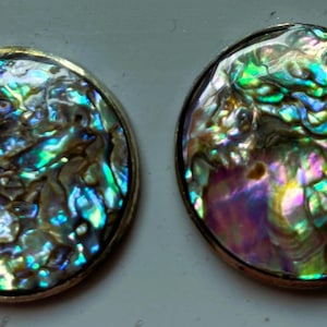 May include: A pair of round earrings with iridescent abalone shells. The shells display a vibrant mix of blues, greens, and purples, set in a silver-toned metal. The earrings are displayed on a light gray surface.