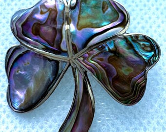 May include: A clover-shaped brooch featuring iridescent abalone shell inlays set within a silver-toned metal frame. The shell displays a spectrum of purple, blue, and green hues with a polished finish, forming a three-leaf clover design.