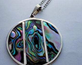 May include: A circular pendant necklace featuring a polished silver-tone frame divided by two vertical silver bars. The inlay consists of iridescent abalone shell displaying swirling patterns of blue, green, purple, and black colors on a chain.