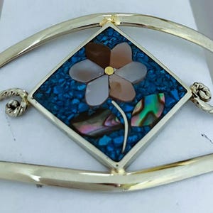 May include: A gold-tone cuff bracelet featuring a central diamond-shaped inlay of crushed blue stone. The inlay displays a flower with light brown and gray petals, a gold center, and iridescent abalone shell leaves and stem.