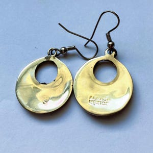 Puede incluir: Un par de pendientes colgantes de metal dorado con un dise&ntilde;o de disco circular y un recorte central. Los pendientes tienen ganchos de alambre curvado. El texto ALPACA MEXICO est&aacute; claramente estampado en la superficie de uno de los discos.