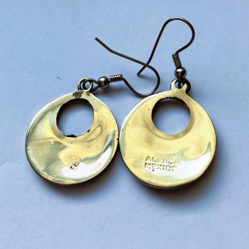 Puede incluir: Un par de pendientes colgantes de metal dorado con un dise&ntilde;o de disco circular y un recorte central. Los pendientes tienen ganchos de alambre curvado. El texto ALPACA MEXICO est&aacute; claramente estampado en la superficie de uno de los discos.
