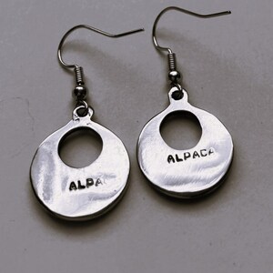 May include: A pair of silver earrings with a circular design. Each earring has a large hole in the center and the word "ALPACA" is engraved on the back.
