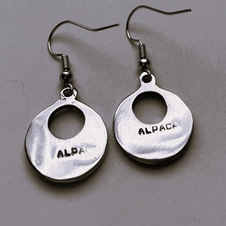 May include: A pair of silver earrings with a circular design. Each earring has a large hole in the center and the word "ALPACA" is engraved on the back.