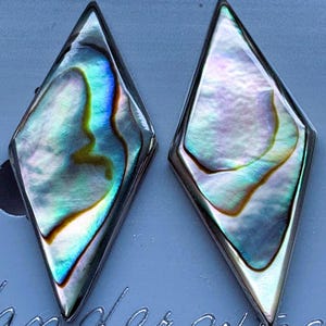 May include: A pair of diamond-shaped earrings featuring iridescent abalone shell inlays set within polished silver-toned metal frames. The shell displays swirling patterns of blue, green, and purple hues, creating a reflective surface that catches the light.