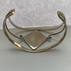 May include: A gold-toned cuff bracelet featuring a central diamond-shaped plaque. The band consists of two parallel wires, with the outer wire having a twisted rope texture. The central plaque is connected to the band by small, coiled metal loops.